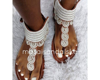 Könnte beinhalten: Weiße Perlensandalen mit braunen Lederriemen und Sohlen. Die Sandalen haben ein einzigartiges Design mit mehreren Perlenlagen und einem dekorativen Muster.