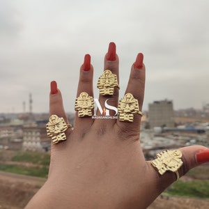 May include: Five gold rings with an Egyptian pharaoh design worn on a hand with red nail polish.