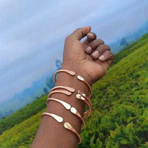 Op de afbeelding: Verschillende koperkleurige open manchetarmbanden worden op een arm gedragen. De armbanden hebben een gehamerde textuur en een eenvoudig, elegant ontwerp. De achtergrond toont een groene heuvel en een bewolkte lucht.