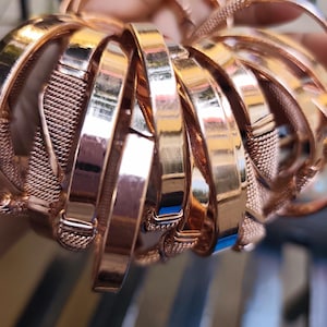 Op de afbeelding: Een verzameling roségouden armbanden. De armbanden variëren in ontwerp, waaronder effen banden en mesh-stijlen. Het gepolijste metaal reflecteert licht, waardoor een warme, glanzende uitstraling ontstaat. De armbanden zijn op elkaar gestapeld en tonen hun verschillende texturen en afwerkingen.