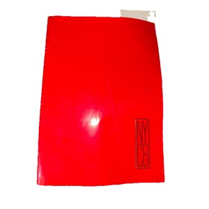 Może przedstawiać: Jasnoczerwony notatnik z literami "NYC B" w czarnym prostokącie w prawym dolnym rogu. Notatnik jest lekko pochylony, z białą etykietą w prawym górnym rogu.