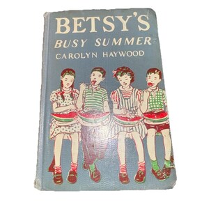 May include: A vintage hardcover book, "Betsy's Busy Summer" by Carolyn Haywood, with a light blue cover. The cover illustration depicts four children enjoying watermelon. The book shows signs of age and use, indicating it's a well-loved, older copy.