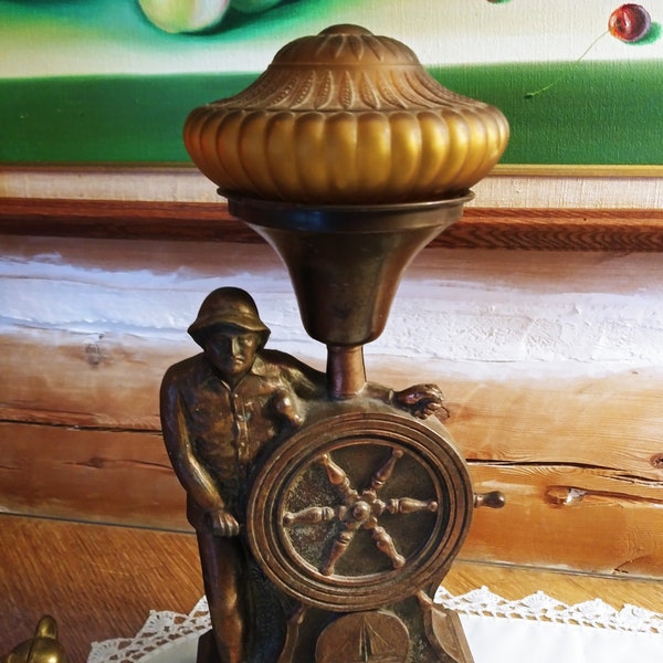 Può includere: Una lampada in ottone vintage con un paralume in vetro smerigliato. La lampada presenta una statuetta di un marinaio che tiene un timone di nave.