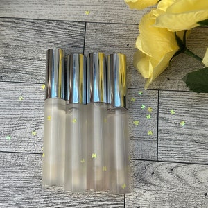 May include: Five clear lip gloss tubes with silver caps and butterfly designs. The tubes are arranged in a row on a light wood surface.