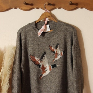 May include: A gray sweater with a crew neck and long sleeves, featuring a design of two flying ducks in shades of brown, white, and black. The sweater is hanging on a wooden hanger with a tag attached.