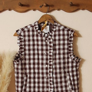 May include: A sleeveless vest in a brown and white gingham pattern, hanging on a wooden hanger. The vest features a ruffled collar, armholes, and hemline, with a button-up front and two front pockets. The vest is made of a quilted fabric.
