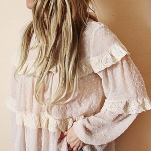 Puede incluir: Una blusa blanca de gasa con un patrón de puntos y ribete de encaje. La blusa tiene un cuello con volantes y mangas largas.