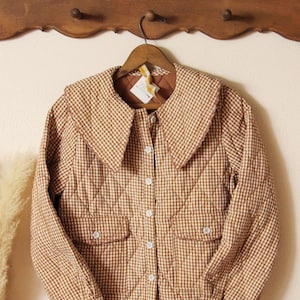 Könnte beinhalten: Eine braun-cremefarbene, gesteppte Jacke mit Vichy-Karo-Muster, großem Kragen und zwei Vordertaschen. Die Jacke hat weiße Knöpfe und hängt an einem Holzbügel. Die Jacke ist im Vintage-Stil.