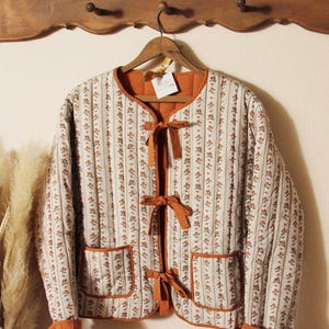 May include: A quilted jacket with a floral and striped pattern in cream, brown, and orange. The jacket has a round neckline, two pockets, and is secured with orange bows. The cuffs and trim are orange.