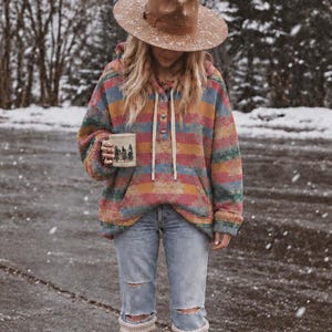 Peut inclure: Un chapeau à larges bords beige, un sweat à capuche avec un motif à rayures horizontales multicolores rouge, jaune et bleu, un jean bleu clair délavé et des bottes en caoutchouc beige avec des revers en tricot blanc, tenant une tasse blanche.