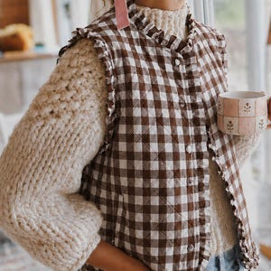 May include: A brown and white gingham quilted vest featuring a ruffled trim along the armholes and front opening, worn over a cream-colored chunky knit sweater. The vest has a button-down front closure and a high neckline with matching ruffles.