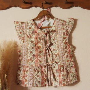 May include: A quilted vest with a floral pattern in cream, red, and pink. The vest has a ruffled hem, tie closures, and small cap sleeves. It hangs on a wooden hanger with a pink ribbon.