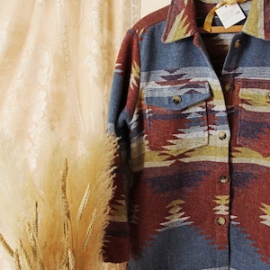 Puede incluir: Una camisa de franela con estampado azul, rojo, amarillo y blanco con cierre de botones y dos bolsillos. La camisa está colgada en una percha de madera.