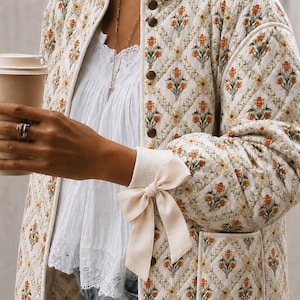 Peut inclure: Une veste matelassée à motif losange floral orange et vert sur fond crème, dotée d'une encolure ronde et d'une fermeture à boutons. Les manches longues présentent des poignets côtelés ornés d'un ruban couleur crème noué en nœud.