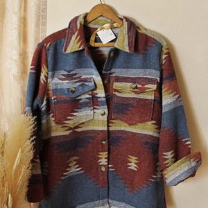 May include: A long-sleeved button-up shirt featuring a multi-coloured geometric pattern in shades of blue, rust red, and mustard yellow. The shirt includes a pointed collar, two chest pockets with button flaps, and a front button closure.