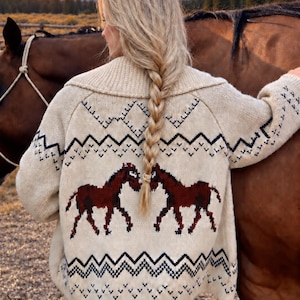 May include: A cream-colored knit cardigan featuring a dark brown horse motif on the back, accented by black and white geometric zigzag patterns along the hem and upper back, complete with a wide ribbed collar and long sleeves.
