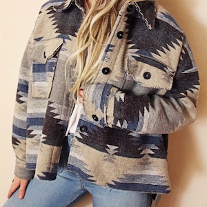 May include: A button-down jacket with a Southwestern-inspired pattern in shades of beige, blue, and gray. The jacket features a collared neckline, two chest pockets, and black buttons. The jacket is worn with blue jeans.