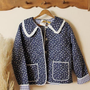 May include: A quilted jacket in navy blue with a white floral pattern. The jacket features a white lace-trimmed collar, white-trimmed pockets, and gingham cuffs. It is buttoned up and hanging on a wooden hanger.