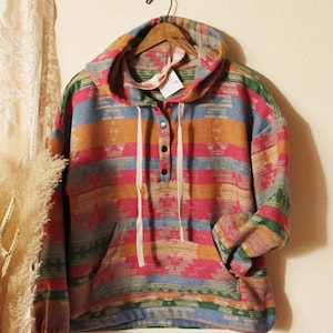 Könnte beinhalten: Ein farbenfroher Kapuzenpullover mit geometrischem Muster in Rosa, Orange, Blau und Grün. Das langärmlige Oberteil hat eine Kängurutasche, einen Kordelzug am Halsausschnitt und drei schwarze Knöpfe. Das Kleidungsstück hängt an einem Holzbügel.