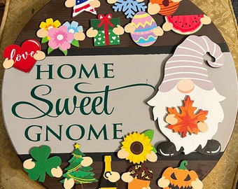Puede incluir: Un letrero redondo de madera con las palabras "Home Sweet Gnome" en letras verdes. El letrero está decorado con varios recortes de fieltro de colores, incluyendo un gnomo, un girasol, una calabaza, un árbol de Navidad, un trébol, un corazón, una estrella, un copo de nieve, un huevo, una sandía, un regalo y flores.