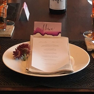 May include: A white oval plate holds a folded menu, a napkin, and a small bouquet of burgundy flowers. A card with the word "Shine" in cursive sits behind the plate. The table setting is on a dark surface.