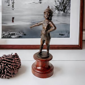 Puede incluir: Figurilla de bronce de un joven con casco y espada, de pie sobre una base de madera.