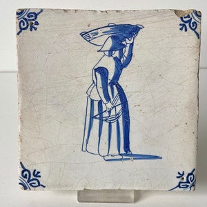 Puede incluir: Un azulejo de Delft azul y blanco que representa a una persona llevando una cesta a la espalda. El azulejo tiene un fondo blanco con diseños florales azules en las esquinas.