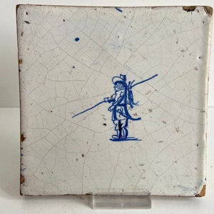 Könnte beinhalten: Eine weiße Keramikfliese mit einem handgemalten Design in Blau und Weiß, das eine Person darstellt, die einen langen Stock auf der Schulter trägt. Die Fliese hat eine craquelée Glasur und ist leicht abgenutzt.