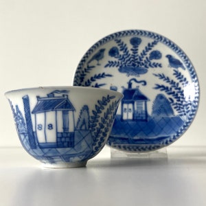 May include: A blue and white porcelain teacup and saucer set with a Chinese-inspired design. The cup and saucer feature a house, trees, and birds.