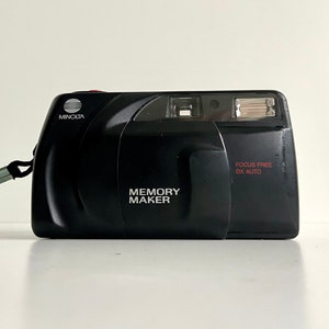 Op de afbeelding: Een zwarte Minolta Memory Maker camera met een zilveren band. De camera heeft een rood "Focus Free DX Auto" label en een zilveren "Memory Maker" label.