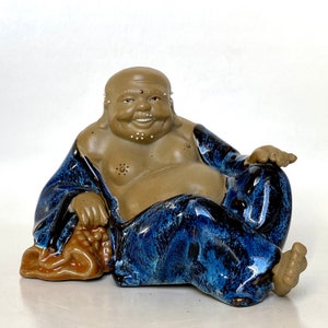 May include: A ceramic figurine of a smiling, chubby Buddha wearing a blue and brown robe. The figurine is sitting with its legs crossed and its hands resting on its lap.
