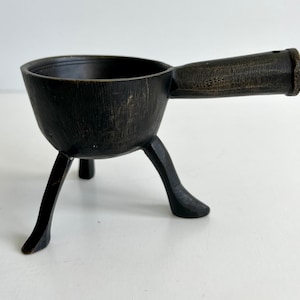 May include: A black metal three-legged bowl with a handle. The bowl has a rustic, aged finish.