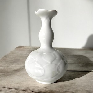 Puede incluir: Un jarrón de cerámica blanca con una base bulbosa y un cuello delgado que se abre en una abertura en forma de pétalo. El jarrón tiene un patrón texturizado en la base y está sobre una superficie de madera.
