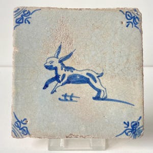May include: Square, off-white ceramic tile with a blue illustration of a running dog. The dog has long ears and a spotted pattern. Blue floral corner designs frame the dog. The tile has a distressed, aged appearance.