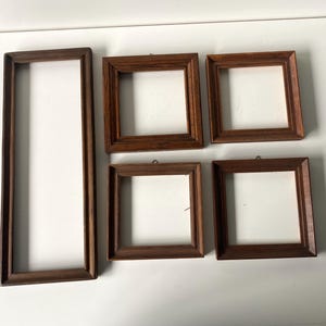 May include: A collection of five wooden picture frames. One is rectangular and vertical, while the others are square. The frames are a dark brown color, with visible wood grain. Each frame has a white interior, ready for photos or artwork.
