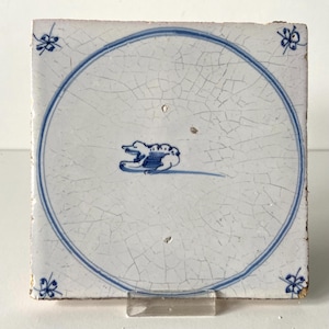 May include: Square, antique ceramic tile with a white background and blue accents. The tile features a blue circle with a stylized animal figure inside. Decorative floral motifs are in each corner. The tile is supported by a clear stand.