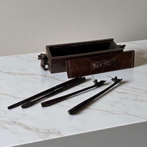 Può includere: Un bruciatore di incenso in legno marrone scuro di forma rettangolare con un cassetto scorrevole. Il cassetto è decorato con un design ornato. Sono inclusi anche diversi strumenti per incenso marrone scuro, tra cui un paio di pinze e due palette per incenso.