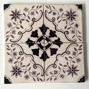 May include: Four square ceramic tiles arranged together, featuring a floral design in dark purple on a cream background. The design includes stylized flowers, leaves, and decorative scrollwork, creating a symmetrical pattern. The tiles appear to be vintage or antique.