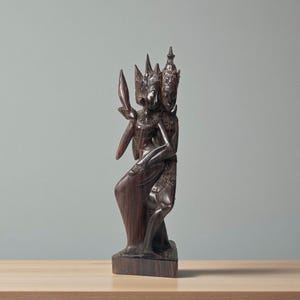 Peut inclure: Une sculpture en bois brun foncé représentant deux personnages, peut-être des danseurs, avec des coiffes ornées et des vêtements détaillés. L'un des personnages tient un objet en forme de feuille. La sculpture est sur une base rectangulaire.