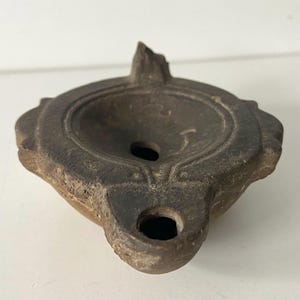 Op de afbeelding: Een donkerbruine klei-olielamp met een rond bassin en een tuit. De lamp heeft een decoratief ontwerp rond de rand en een klein gaatje in het midden van het bassin.
