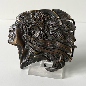 May include: A bronze belt buckle featuring a detailed sculpture of an American Indian chief wearing a headdress with feathers.