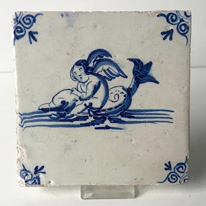 Puede incluir: Azulejo cerámico cuadrado con diseño azul y blanco. La imagen presenta un querubín montando una criatura marina, con detalles decorativos en las esquinas. El azulejo tiene un aspecto envejecido.