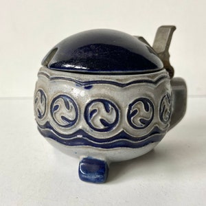 Puede incluir: Un tarro de cerámica azul y blanco con tapa y asa de peltre. El tarro tiene un diseño decorativo de círculos con un patrón de hojas estilizadas. El tarro tiene tres patas.