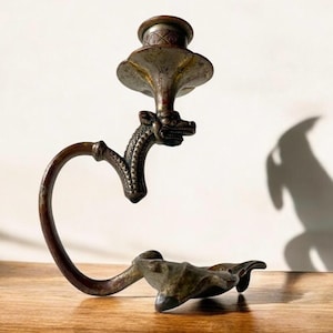 May include: A bronze candlestick with a dragon design. The candlestick has a curved arm and a round base. The dragon's head is facing forward and the candlestick is in a rustic style.