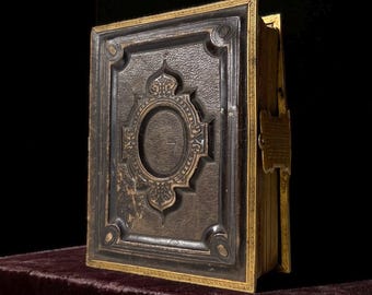 Puede incluir: Un libro antiguo con una cubierta oscura y texturizada y detalles dorados. La cubierta presenta un diseño en relieve con un centro ovalado y detalles ornamentados. Los bordes de las páginas están dorados y un cierre dorado asegura el libro.