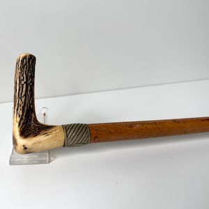 Puede incluir: Un bastón con un mango en forma de L hecho de asta texturizada en tonos marrón y crema. El mango se conecta a un eje de madera lisa de color marrón claro mediante un collar decorativo de metal plateado estriado que rodea la unión.