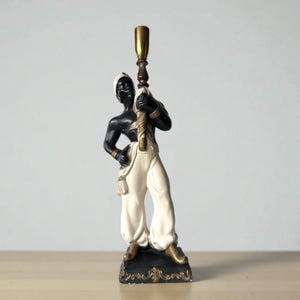 May include: A vintage figurine of a standing figure in white trousers and a turban, holding a tall, ornate candle holder. The figure is black with gold accents, and the base is black with gold detailing. A decorative piece.
