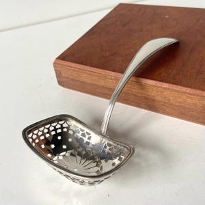 May include: A silver-colored serving ladle with a decorative pierced bowl featuring star and floral patterns. The handle curves gracefully, and the ladle rests on a wooden surface. The ladle is designed for serving.