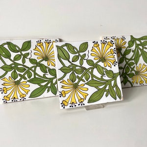 Peut inclure: Trois carreaux de céramique carrés avec un motif floral. Les carreaux présentent un fond blanc avec des vignes vertes et des fleurs jaunes.