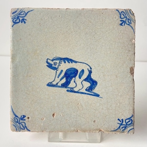 May include: Square ceramic tile with a light blue background and a blue bear illustration. The bear is depicted walking on a blue line. Decorative blue floral corner accents are present.